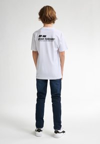 Bílý bavlněný tričko s černým logem "GRAN TURISMO" na zadní straně, kombinované s tmavě modrými džíny a bílými teniskami s černými akcenty.