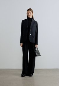 Zwart op maat gemaakte blazer met een enkele knoop, gedragen over een zwarte coltrui en bijpassende broek met wijde pijpen. Houdt een kleine zwarte handtas vast.