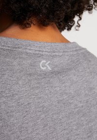 T-shirt en coton gris avec col rond et manches courtes. Présente un petit logo blanc au dos, près de l'encolure. La texture semble douce.