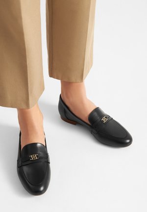 Mocassini neri in pelle con logo a accento dorato, caratterizzati da punta arrotondata e suola piatta. Abbinati a pantaloni su misura color cognac.