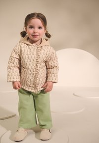 Veste matelassée beige avec motif floral, capuche et boutons-pression, associée à un pantalon vert clair et des chaussures à enfiler couleur crème. Design simple.