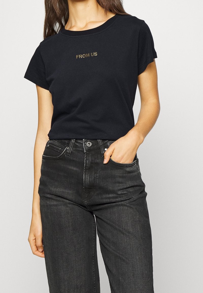 Svart bomullströja med korta ärmar, med guldtexten "FROM US". Kombinerad med mörkgrå jeans som framhäver en klassisk passform och enkel design.