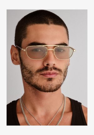 Kuldraamides prillid heledate toonidega läätsede, ruudukujulise kuju, minimaalse disaini ja õhukeste templitega, kantud lühema juustega inimesel.