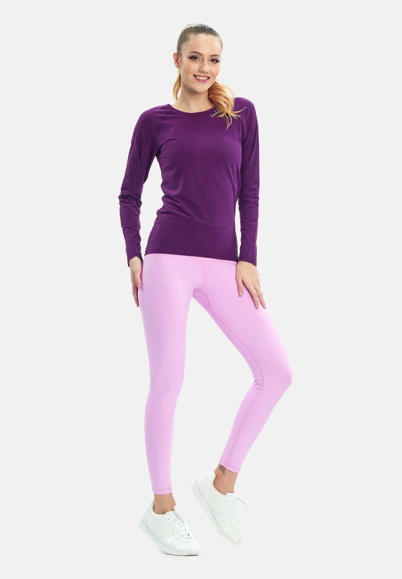 Ljubičasta majica dugih rukava u kombinaciji sa svijetlopinkastim tajicama. Outfit je izrađen od glatkog, rastezljivog materijala. Bijeli tenisice zaokružuju izgled.