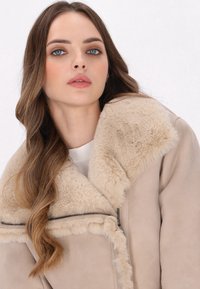 Veste en suédine beige avec un col en fausse fourrure, dotée d'une fermeture éclair sur le devant et d'un tissu texturé, mettant en avant une apparence douce et chaleureuse.