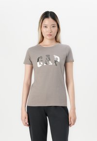 Woman wearing a gray short-sleeve T-shirt with "GAP" in large metallic letters and black pants, standing against a plain white background.