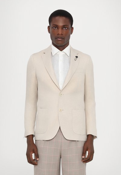Beige Blazer mit taillierter Passform, zwei vorderen Taschen und einem Revers-Pin, getragen über einem weißen Hemd. Kombiniert mit grauen karierter Hose.