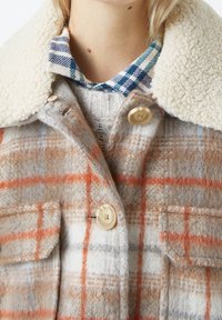 Giacca di lana a quadri con motivi beige e arancione, caratterizzata da un colletto in crema e morbido e chiusure con bottoni, abbinata a una camicia a scacchi blu sottostante.