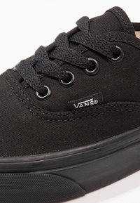 Crne platnene tenisice s okruglim prstom, ravnim potplatom i crnim vezicama. Ima metalne patentne kopče i Vans oznaku na bočnoj strani.