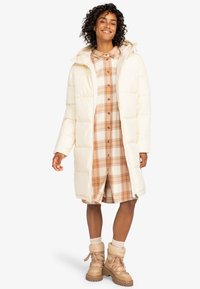 Doudoune crème avec capuche, présentant un design matelassé. En dessous, une robe chemise à carreaux crème, orange et marron avec des boutons. Bottes beiges.