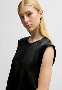 Top nero senza maniche in tessuto liscio con scollo rotondo e design delle spalle strutturato, caratterizzato da una vestibilità rilassata e uno stile minimalista.