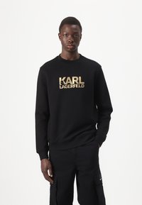 Sudadera negra con el logo dorado de "KARL LAGERFELD". Diseño de cuello redondo, mangas largas, tela suave, puños y dobladillo acanalados. Combinada con pantalones cargo negros.