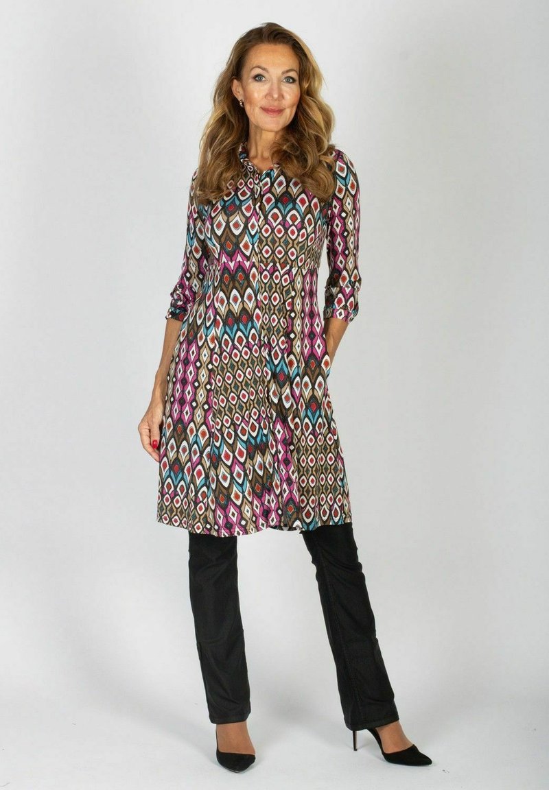 Kobieta z długimi falującymi włosami, ubrana w kolorową sukienkę w geometryczny wzór na czarnych spodniach i czarnych szpilkach, stojąca na jednolitym tle.