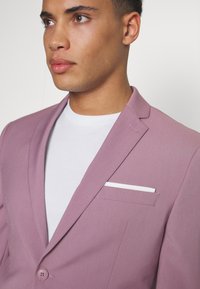 Svijetlopurple blejzer napravljen od glatkog materijala, s jednim dugmetom za zatvaranje, oštrim reverima i bijelom maramicom u bočnoj džepu.