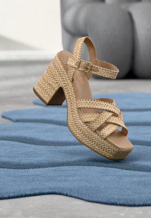 Gewebte beige Plateausandale mit klobigem Absatz und Knöchelriemen mit Schnalle, liegend auf strukturiertem blauem Teppich mit grauem Hintergrund.