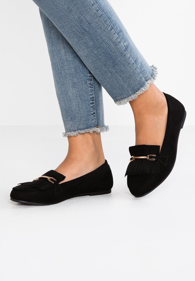 Jambes portant un jean bleu clair à ourlet effiloché et des mocassins en daim noir avec franges et détail de boucle dorée sur un sol blanc.
