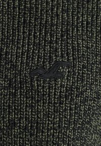 Stickad tröja i mörkgrön med texturerad ribbning. Har en svart broderad logotyp på framsidan som visar på särskilt hantverk.
