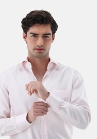 Helles rotes Langarmhemd mit einem Button-Down-Kragen, einer Brusttasche und strukturiertem Stoff. Das Model stellt den Manschettenverschluss ein, während es nach vorne schaut.