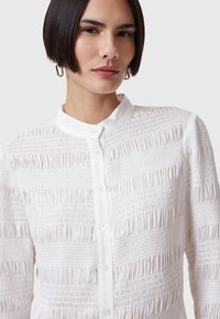 Chemise blanche à boutons avec un design texturé et froncé. Caractéristiques : col montant et fermeture avant à petits boutons. Tissu lisse.