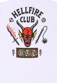 Bílý tričko s grafickým potiskem zobrazující červenou lebku s rohy, plameny, meči a textem "HELLFIRE CLUB" v různých barvách a stylech.