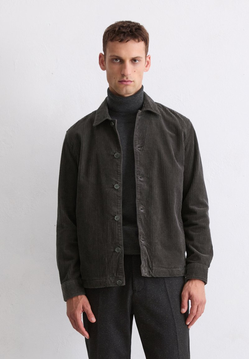 Dark green corduroy jacket with a button-up front, a pointed collar, and long sleeves. Worn over a fitted gray turtleneck. Textured fabric.