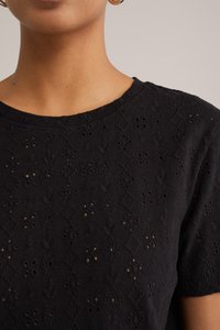 Melnā blūze ar ziedu izšuvumiem un izgriezumiem, ar apaļu kakla izgriezumu un īsām piedurknēm. Tekstūrēts audums uzlabo vizuālo pievilcību.