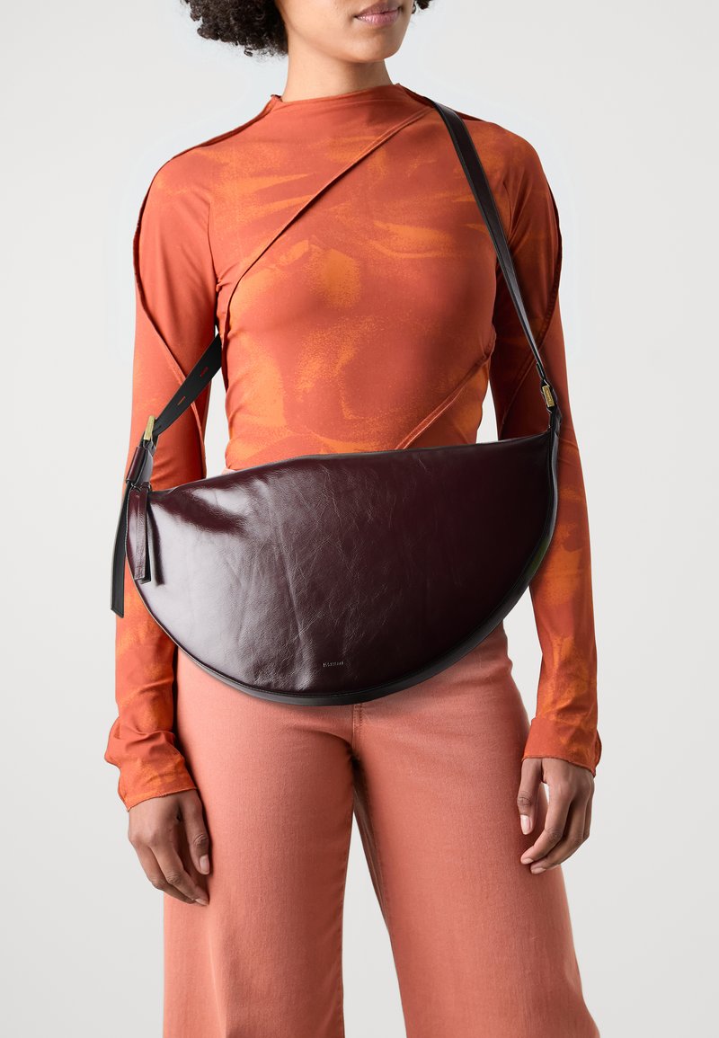Mørk burgunder skinn crossbody-veske med en semi-sirkelform, svart kant og glidelås-lukking. Modellen har på seg en oransje langermet topp.