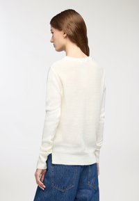Maglione cremoso chiaro con scollo rotondo, maniche lunghe e polsini a coste. Morbido tessuto in maglia. Abbinato a jeans blu larghi.