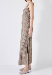 Robe longue sans manches en tissu beige texturé avec des rayures verticales ; offre une fente latérale haute et une coupe décontractée. Portée avec des ballerines scintillantes.