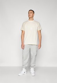 Beige grafisk t-skjorte med "NEW YORK YANKEES" trykk, kombinert med grå cargo joggebukser og hvite sneakers. Uformell sporty bekledning.