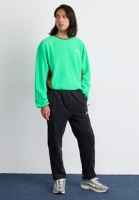 Grüner Fleece-Sweatshirt mit braunen Seitenteilen, kombiniert mit schwarzen Fleece-Hosen. Auffälliges Logodetail auf dem Sweatshirt und den Hosen.