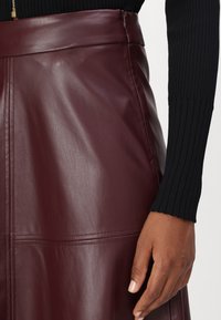 Detail osoby nosiacej bordovú koženú sukňu s prešívanými detailmi a čierny rebrovaný top s dlhým rukávom, ruka oddychuje pri boku.