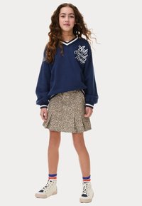 Sweatshirt marine avec des lettres blanches, associé à une jupe plissée à imprimé léopard. Le mannequin porte des baskets montantes crème avec des chaussettes rayées.
