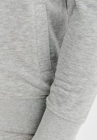 Materiał szarej bluzy dresowej o miękkiej fakturze. Posiada boczną kieszeń i dopasowane mankiety. Projekt podkreśla wygodę i swobodny styl.