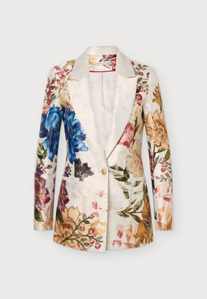 Femme aux longs cheveux blonds, portant une robe blazer fleurie et des bottes en cuir marron montantes jusqu'aux genoux, debout devant un fond clair uni.