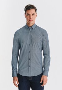 Niebieska koszula z długim rękawem z wzorem, z kołnierzykiem zapinanym na guziki, z geometrycznym wzorem, dopasowana, z ciemnymi guzikami. Noszona z ciemnymi dżinsami.