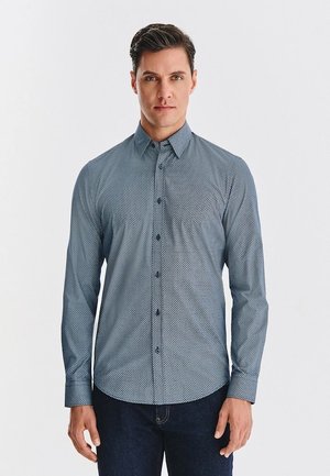 Niebieska koszula z długim rękawem z wzorem, z kołnierzykiem zapinanym na guziki, z geometrycznym wzorem, dopasowana, z ciemnymi guzikami. Noszona z ciemnymi dżinsami.