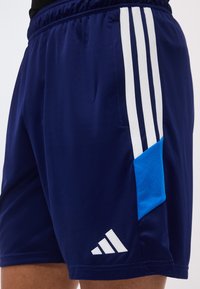 Σκούρο μπλε αθλητικό σορτς με λευκές κάθετες ρίγες, μπλε δίχτυ πλαϊνό κομμάτι και λευκό λογότυπο Adidas κοντά στο τελείωμα.