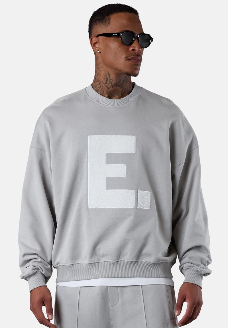 Sweat-shirt gris à col rond avec une grande lettre "E" blanche en relief sur le devant. Présente une coupe décontractée et des bordures côtelées à l'ourlet et aux poignets.