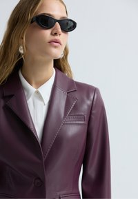 Blazer en cuir bordeaux ajusté avec des revers crantés, des coutures détaillées et une fermeture à bouton, associé à une chemise blanche et des lunettes de soleil noires.