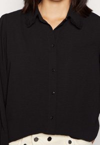 Chemise noire à boutons, en tissu texturé, avec un col en dentelle et des manches longues. Se ferme avec des boutons noirs, portée sur un pantalon de couleur claire.