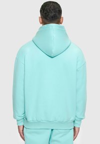 Helle aquafarbene, übergroße Kapuzenjacke mit Kordelzug, gerippten Bündchen und Bund. Glatte Textur mit entspannter Passform, von hinten betrachtet.