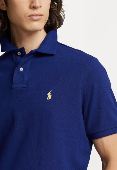 Temno modra polo majica iz teksturiranega bombaža, z zloženim ovratnikom in rumenim izvezenim logotipom na prsih. Dva gumba za zapenjanje.