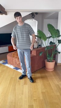 Fiatal férfi szürke csíkos ingben és világoskék nadrágban áll a nappaliban, egy barna kanapé mellett, és egy nagy cserépben lévő növény közelében.