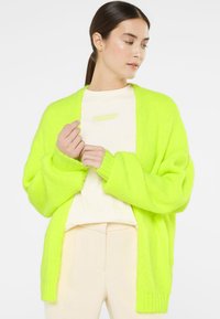 Neon green oversized cardigan, knitted texture, open front, dropped shoulders, paired with a cream top and light-colored pants.