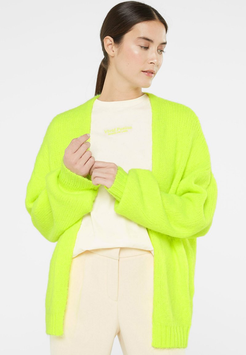 Neon green oversized cardigan, knitted texture, open front, dropped shoulders, paired with a cream top and light-colored pants.