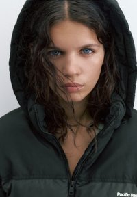 Mladá žena s modrými očami a kučeravými vlasmi nosí čiernu kapucňu so zapnutým zipsom a pozerá priamo pred seba s neutrálnym výrazom.