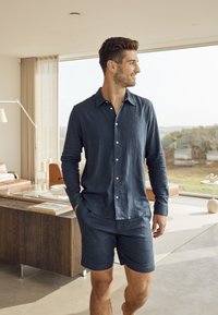 Marineblaues Leinenhemd mit langen Ärmeln und weißen Knöpfen, kombiniert mit passenden Shorts. Glatte Textur; lässig in einem hellen Innenraum getragen.