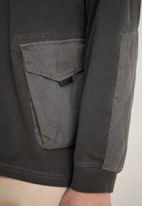 Gray long-sleeve shirt featuring a textured pocket with a flap, ribbed cuffs, and a subtle quilted pattern on the pocket material.