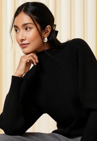 Schwarzer, gerippter Pullover mit hohem Kragen und langen Ärmeln. Der Stoff hat eine strukturierte Oberfläche. Ein Paar silberne Ohrringe sind sichtbar. Die Hand des Modells ruht auf dem Kinn.